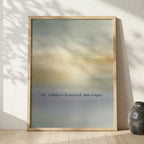A framed art print leaning against a textured white wall on a light wood floor, next to a small dark ceramic vase. The print features an abstract soft gradient background in muted blue, beige, and yellow tones with the text 'My stillness is sacred, not empty' in a small black serif font.