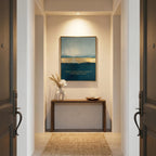 A framed abstract landscape art print with blue, beige, and gold tones hangs on a beige wall in a hallway above a wooden console table with a vase of dried grass and a small bowl. The hallway is framed symmetrically by dark wood doors on both sides.