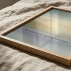 A square framed art print with a light wood frame lies on a textured beige linen surface. The print features a soft gradient background with the text 'Silence is sacred, not empty' in a thin dark font.
