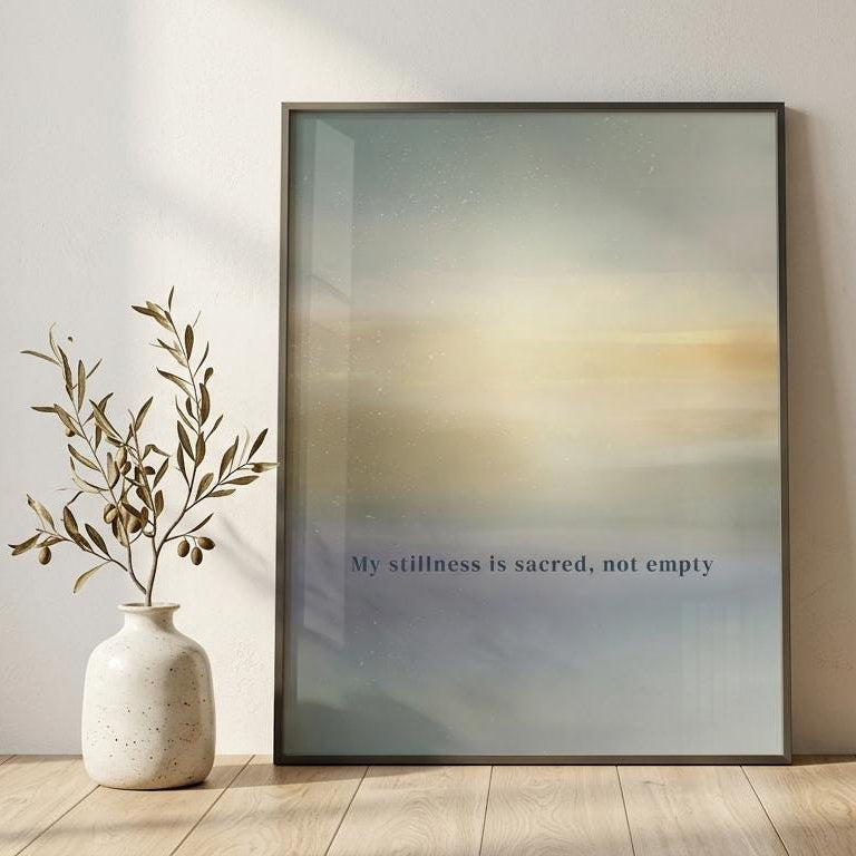 A framed minimalist art print leaning against a light beige wall on a wooden floor, next to a white ceramic vase with dried branches. The print features a soft gradient background transitioning from blue-gray to warm beige and yellow, with the text 'My stillness is sacred, not empty' printed near the bottom.