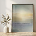 A framed minimalist art print leaning against a light beige wall on a wooden floor, next to a white ceramic vase with dried branches. The print features a soft gradient background transitioning from blue-gray to warm beige and yellow, with the text 'My stillness is sacred, not empty' printed near the bottom.
