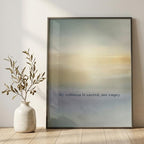 A framed minimalist art print leaning against a light beige wall on a wooden floor, next to a white ceramic vase with dried branches. The print features a soft gradient background transitioning from blue-gray to warm beige and yellow, with the text 'My stillness is sacred, not empty' printed near the bottom.