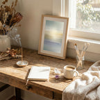 A framed minimalist art print with a soft blue and beige abstract design leans on a rustic wooden table in a sunlit room. The scene includes a notebook, a mug of coffee, a glass jar of paintbrushes, dried flowers, and a textured throw blanket.