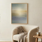 A framed abstract art print with soft blue, beige, and yellow tones hangs on a cream wall in a bright, modern living room setting with a beige armchair, wooden side table, and potted plant.