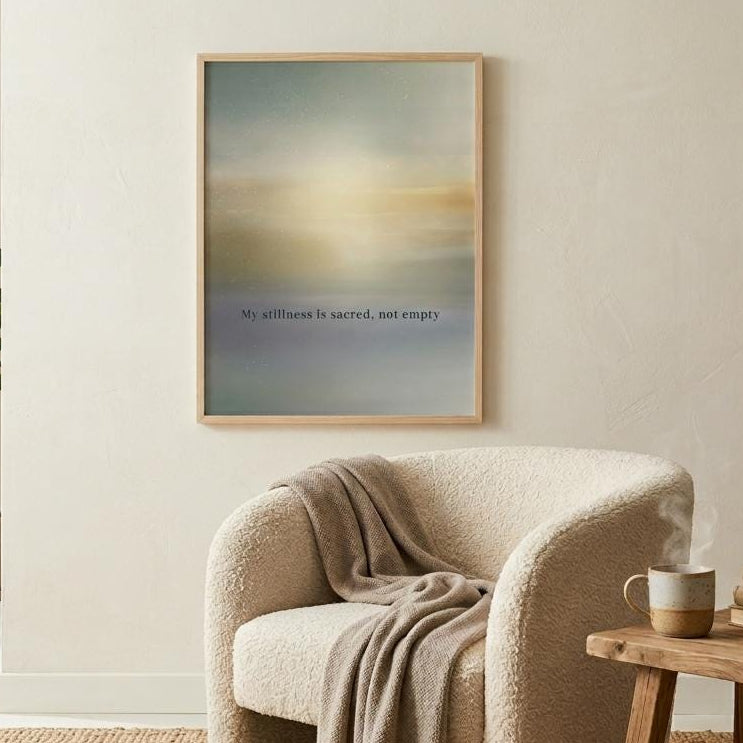 A framed abstract art print with soft blue, beige, and yellow tones hangs on a cream wall in a bright, modern living room setting with a beige armchair, wooden side table, and potted plant.