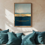 A framed abstract art print hangs on a textured beige wall above a sofa with teal pillows. The artwork features a blue and gold landscape with a quote in gold text at the bottom.